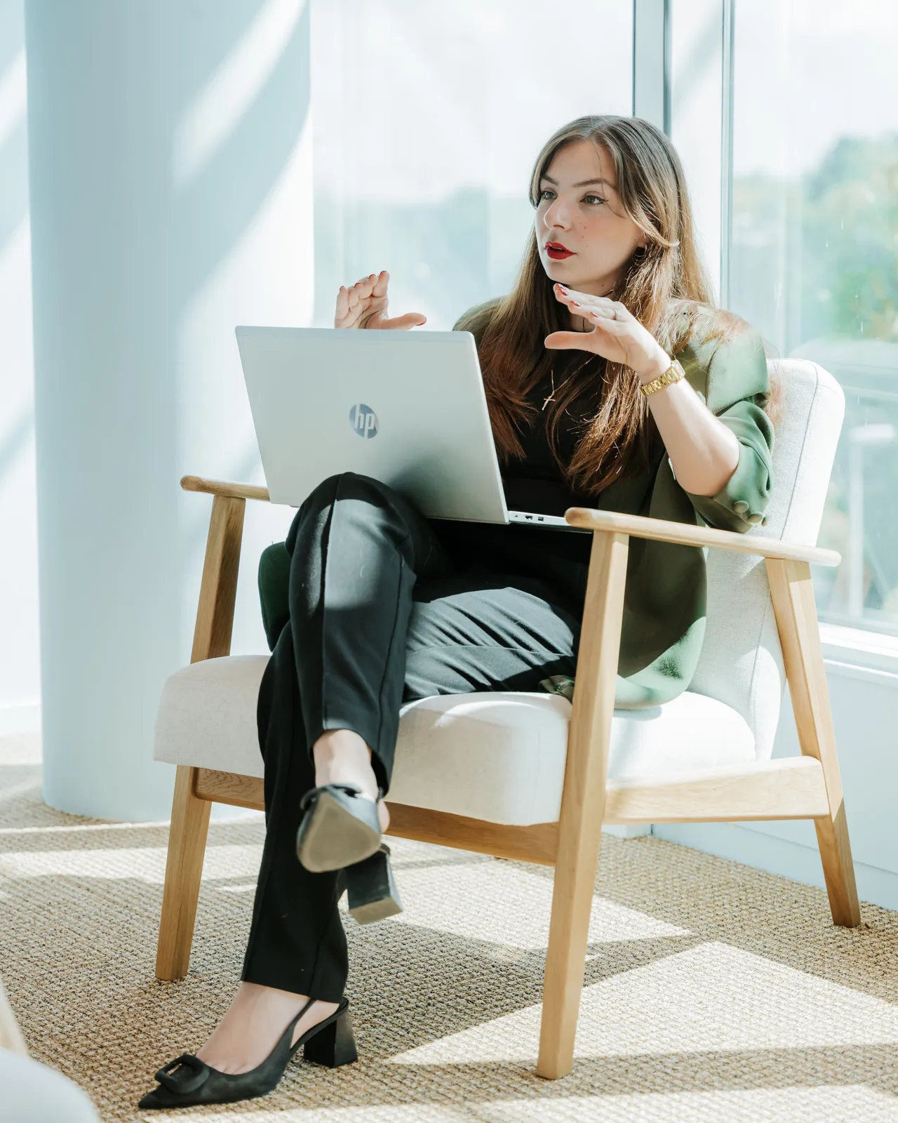 Woman sitting in a chair by a window, gesturing while using a laptop, wearing a green blazer and black pants.