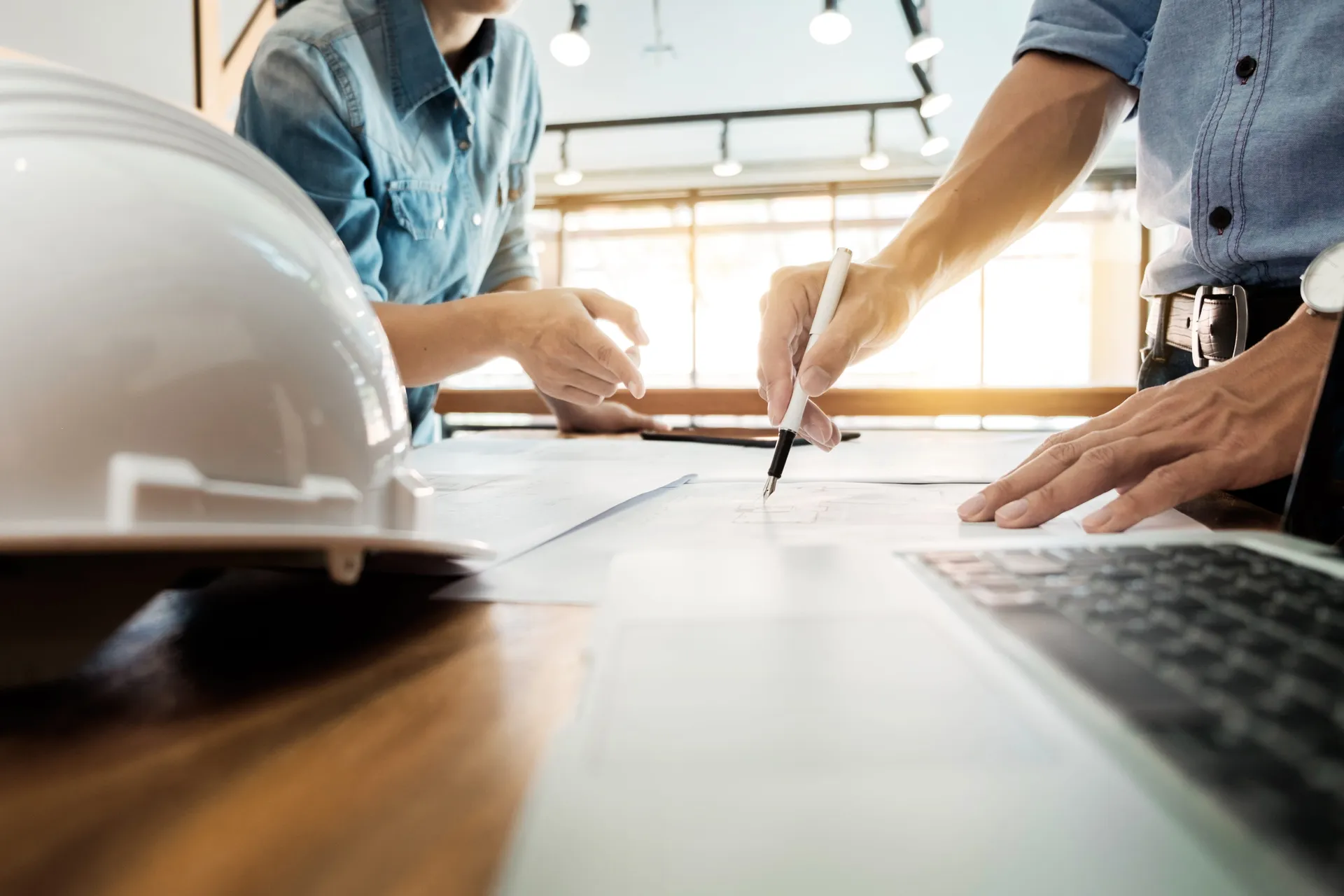 Two engineers review blueprints on a desk with a laptop and white safety helmet in a bright office.