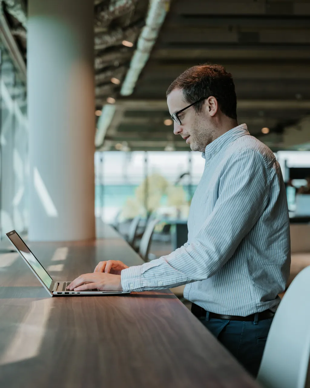 Man typing on a laptop at a standing desk in a modern office with large windows and natural light.