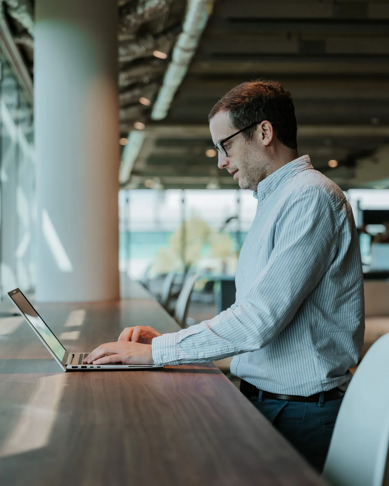 Man typing on a laptop at a standing desk in a modern office with large windows and natural light.