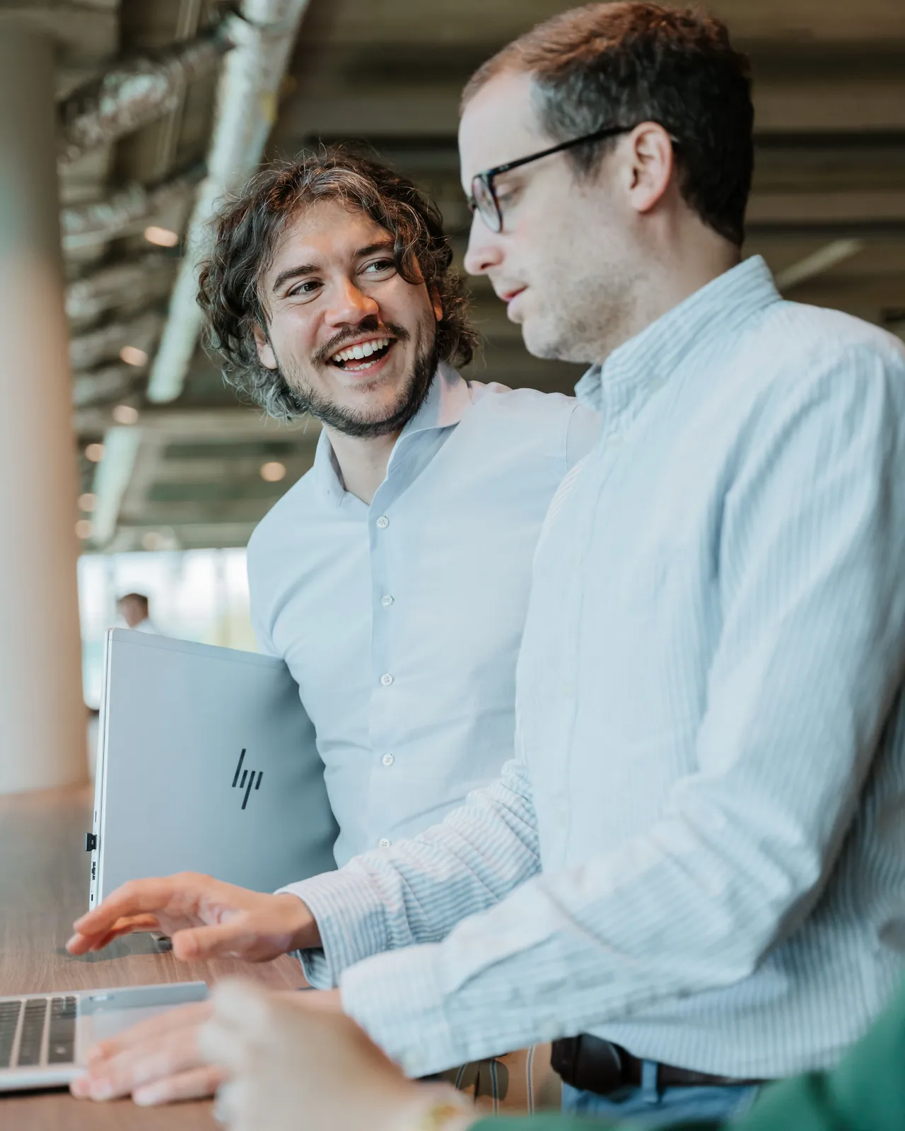 Two men in casual shirts, one holding an HP laptop, engage in a friendly conversation in a modern office setting.