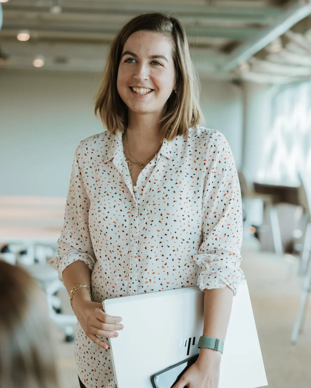 Smiling woman in a polka dot shirt holds a laptop and phone, standing in a bright room with modern decor.