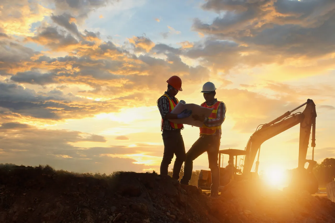 Two construction workers in hard hats and reflective vests reviewing plans beside an excavator on a construction site at sunset.