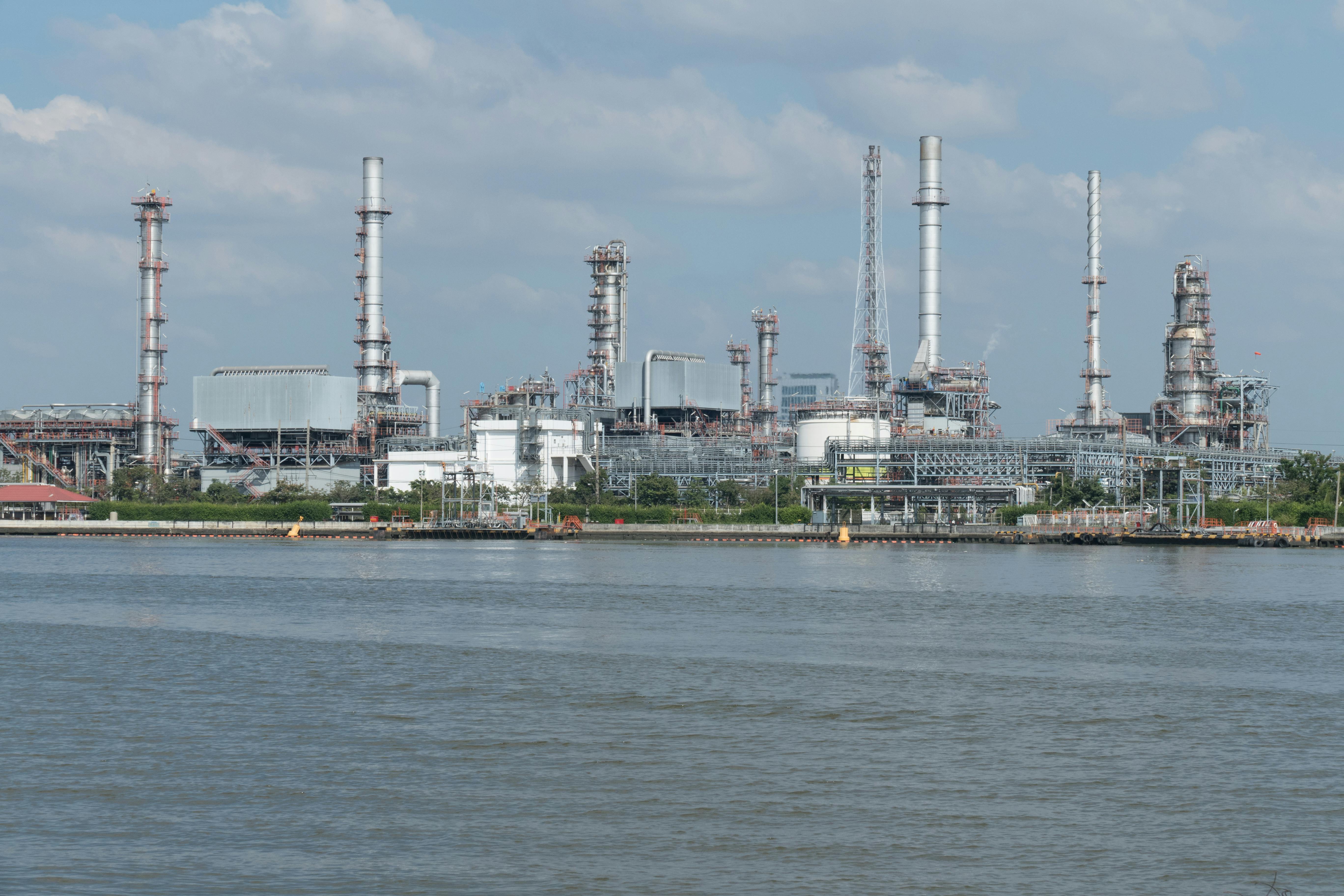 Industrial refinery complex with tall chimneys and metal structures by a body of water under a cloudy sky.