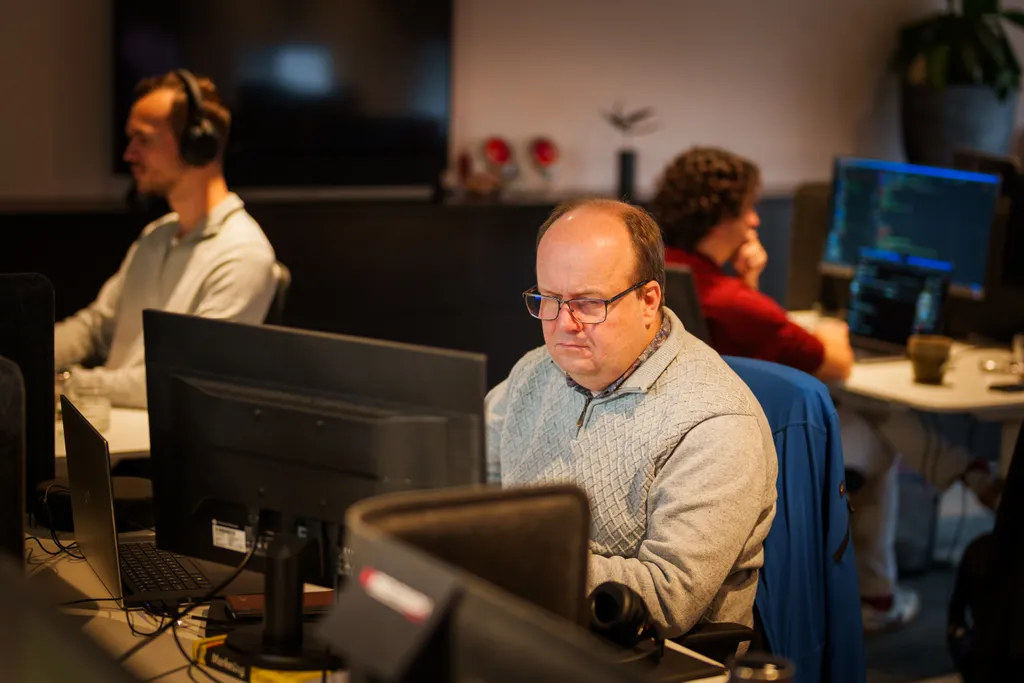 Drie mensen werken aan computerbureaus in een kantooromgeving. De man op de voorgrond, met een bril en een trui, richt zich op zijn monitor. Twee anderen zijn zichtbaar, een met een koptelefoon en een ander die typt, met schermen waarop code wordt weergegeven.