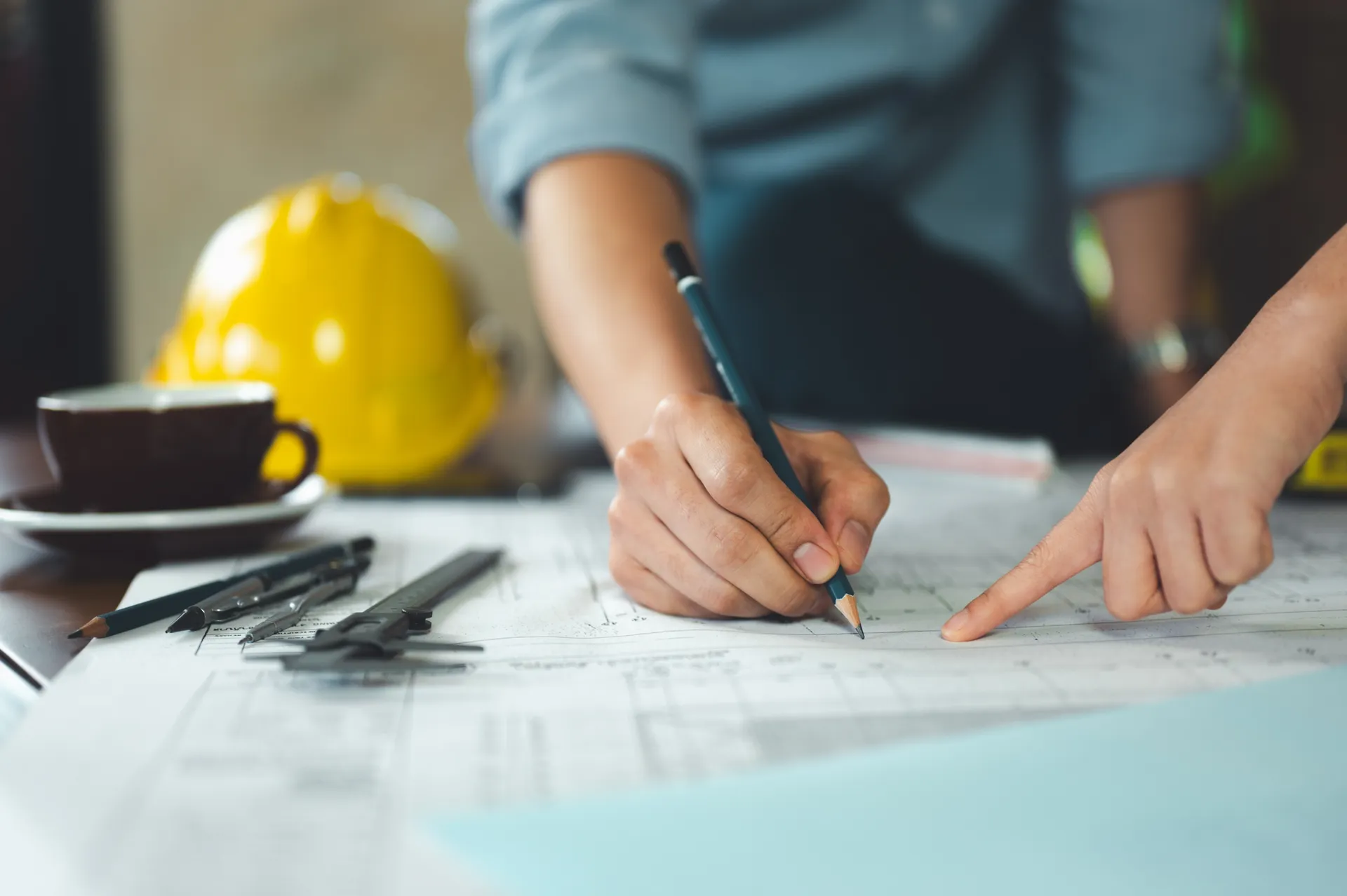 Close-up van handen die met potlood een technische tekening aanpassen op een bureau met gereedschap en helm.