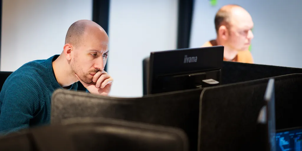 Twee mannen zitten aan aparte computerwerkplekken in een kantooromgeving. De man op de voorgrond is geconcentreerd, met zijn kin op zijn hand, terwijl hij naar een scherm kijkt. Beiden zijn omgeven door donkere verdelers, wat een geconcentreerde werkruimte creëert.