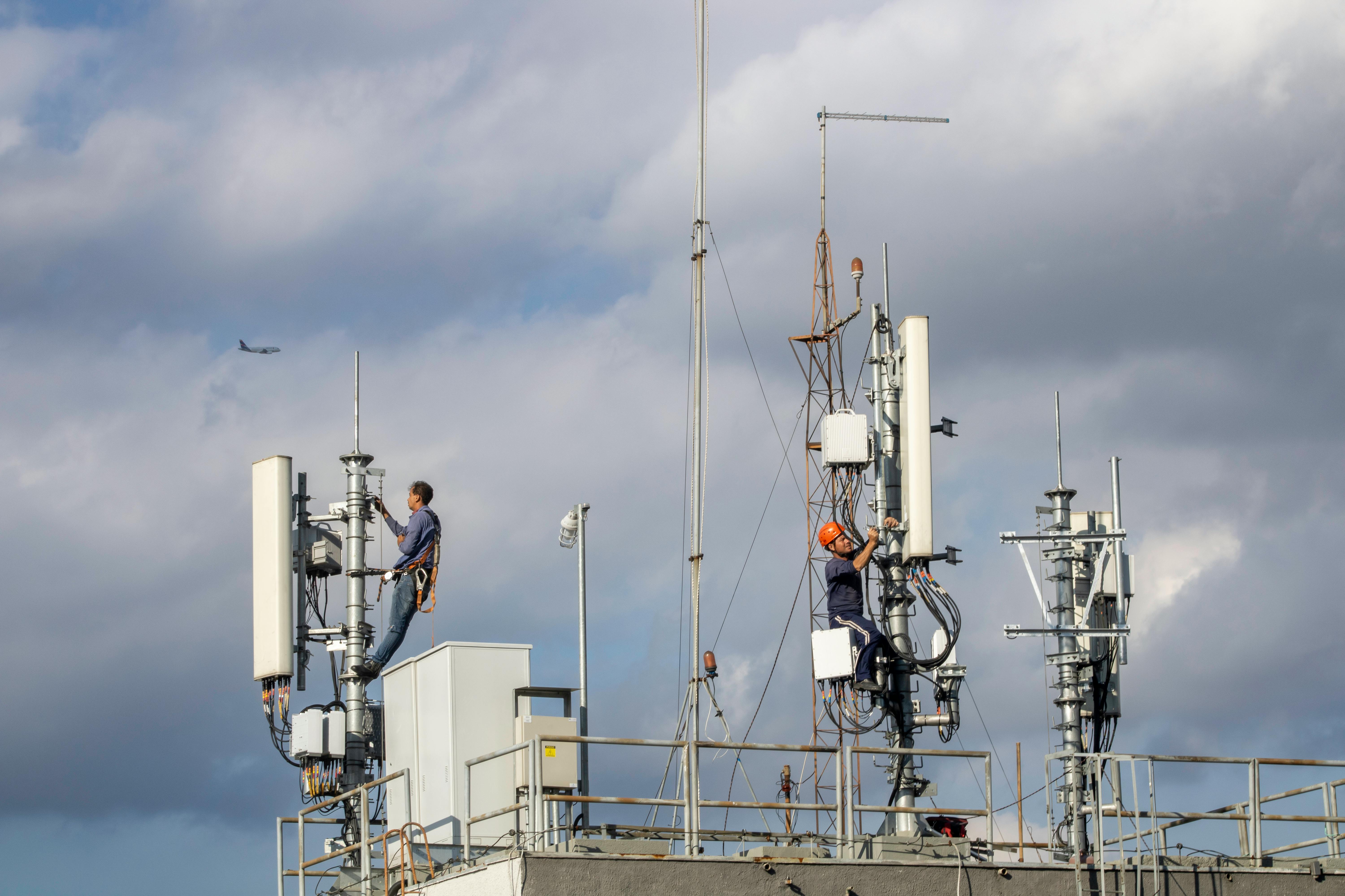 Two workers in safety gear maintain antennas on a rooftop. Cloudy sky and distant airplane in the background.