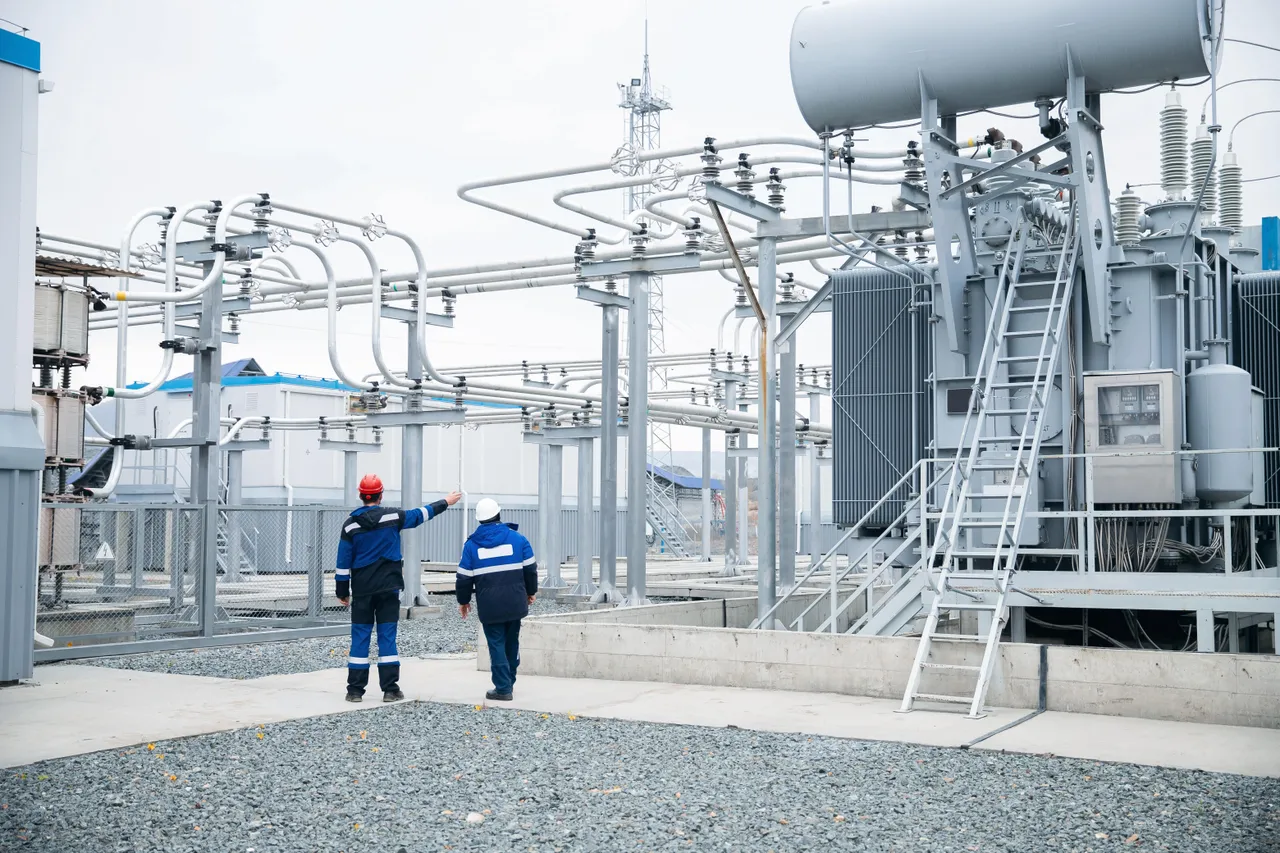 Two utility workers in hard hats inspect a large transformer and overhead buswork at an electrical substation.