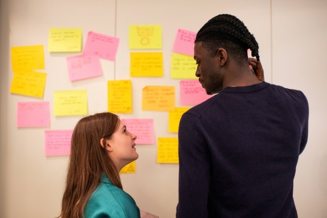 Two people stand in front of a wall covered with colorful sticky notes, engaged in conversation.