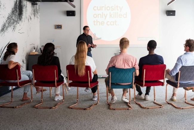 A man presents to a diverse group seated in a modern room with a screen displaying the phrase 
