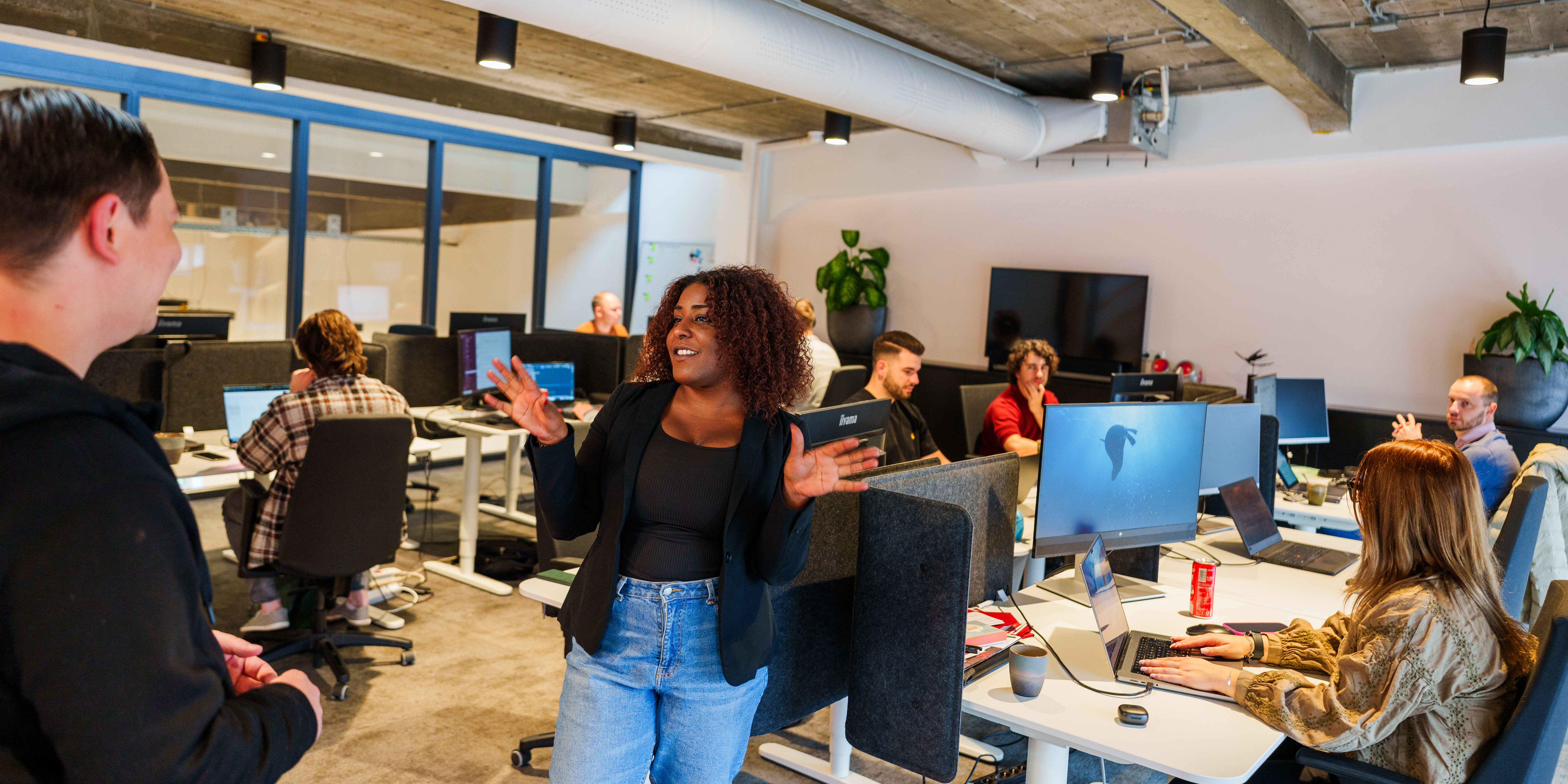 Een levendige kantooromgeving met diverse werknemers die aan computerbureaus werken. Een vrouw staat en praat geanimeerd met een collega, terwijl anderen zich op hun schermen concentreren. De kamer is modern met zichtbare plafonds en planten, wat een collaboratieve sfeer creëert.