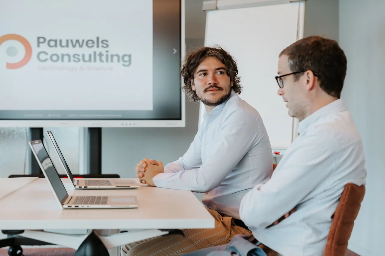 Two men sit at a table with laptops, engaged in discussion. A screen displays "Pauwels Consulting Technology & Science" in the background.