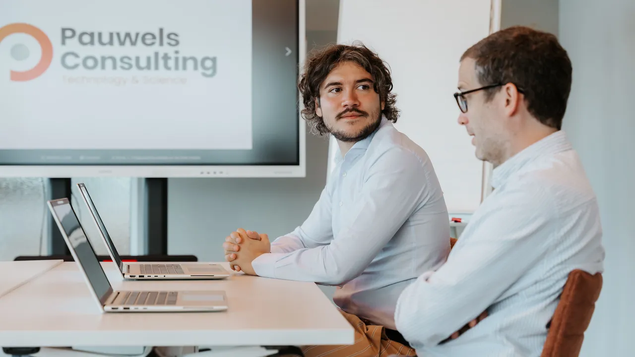 Two men sit at a table with laptops, engaged in discussion. A screen displays "Pauwels Consulting Technology & Science" in the background.
