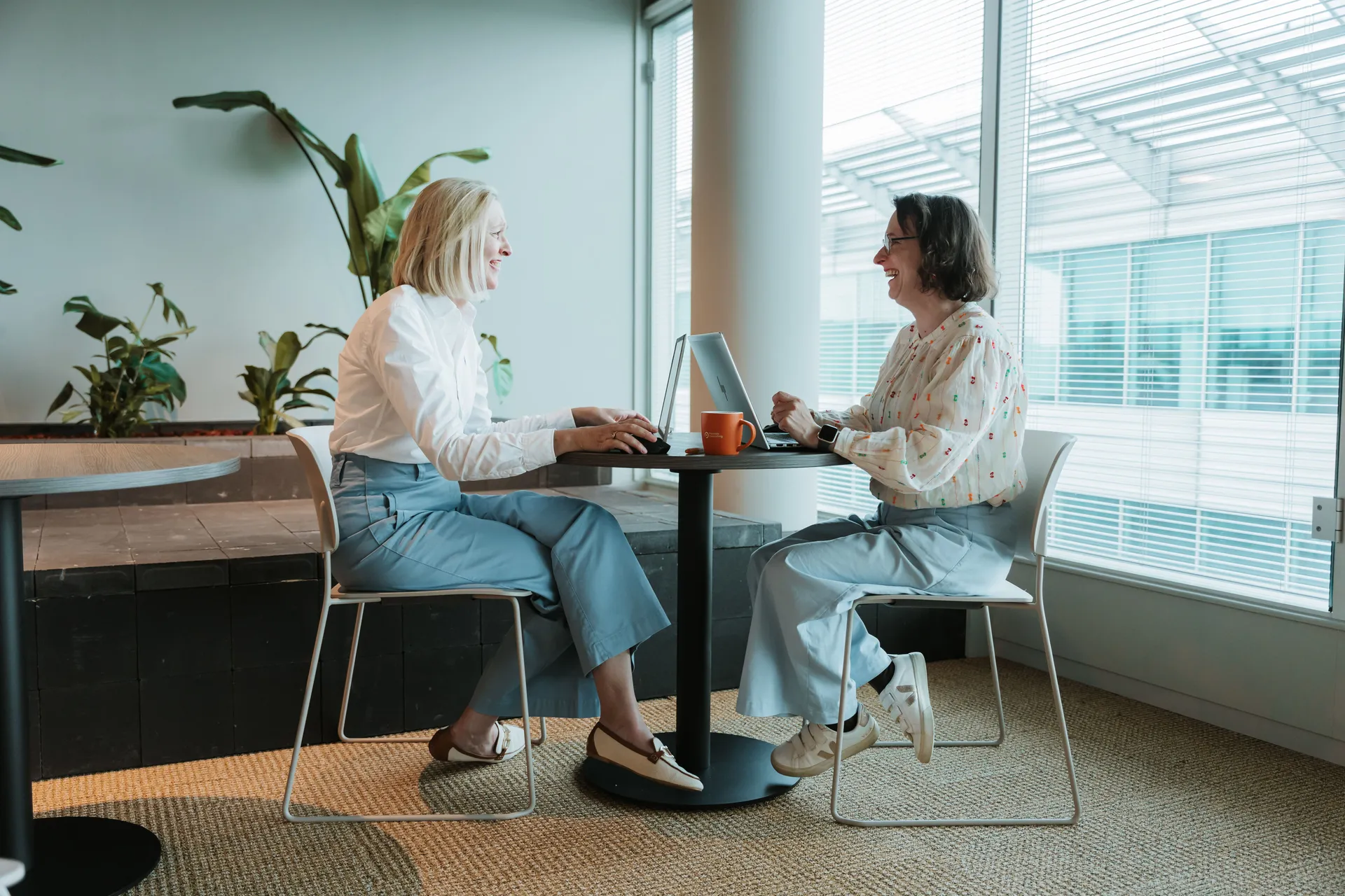 Two women sit at a small round table in an office, engaged in conversation, with laptops and coffee mugs. Plants and large windows are in the background.