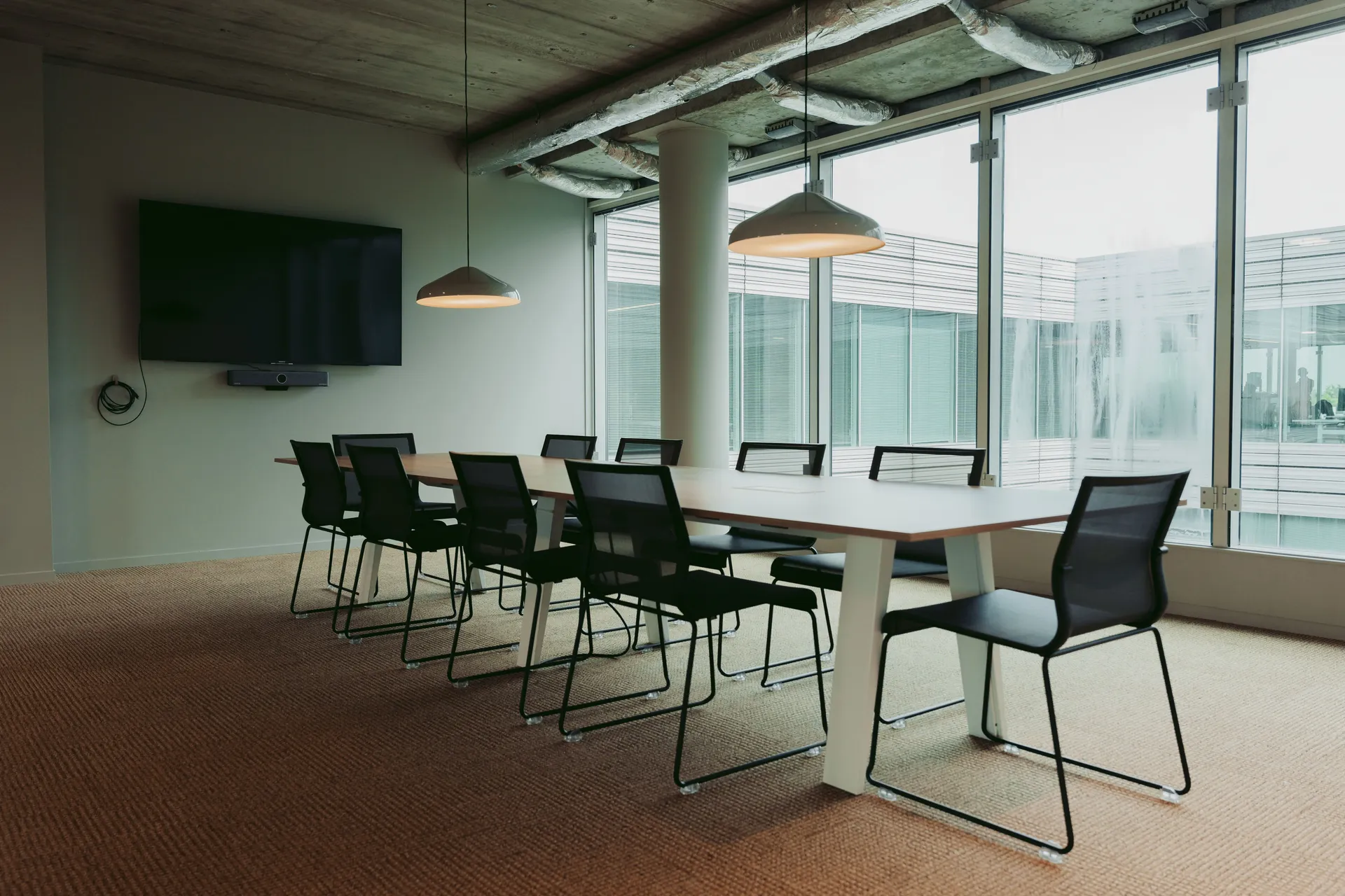 Modern conference room with a long table, black chairs, ceiling lights, a wall-mounted TV, and large windows with a view.