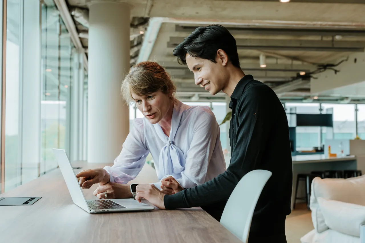 Two people in a modern office setting, collaborating and looking at a laptop on a wooden table near large windows.