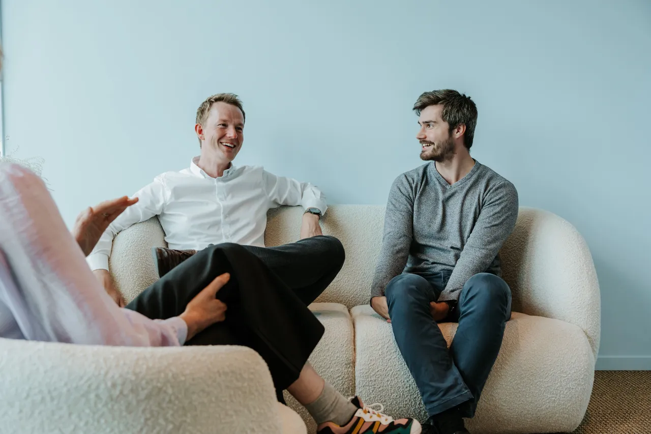 Three people sitting on a sofa in a casual setting, engaged in a lively conversation, with two men smiling at each other.