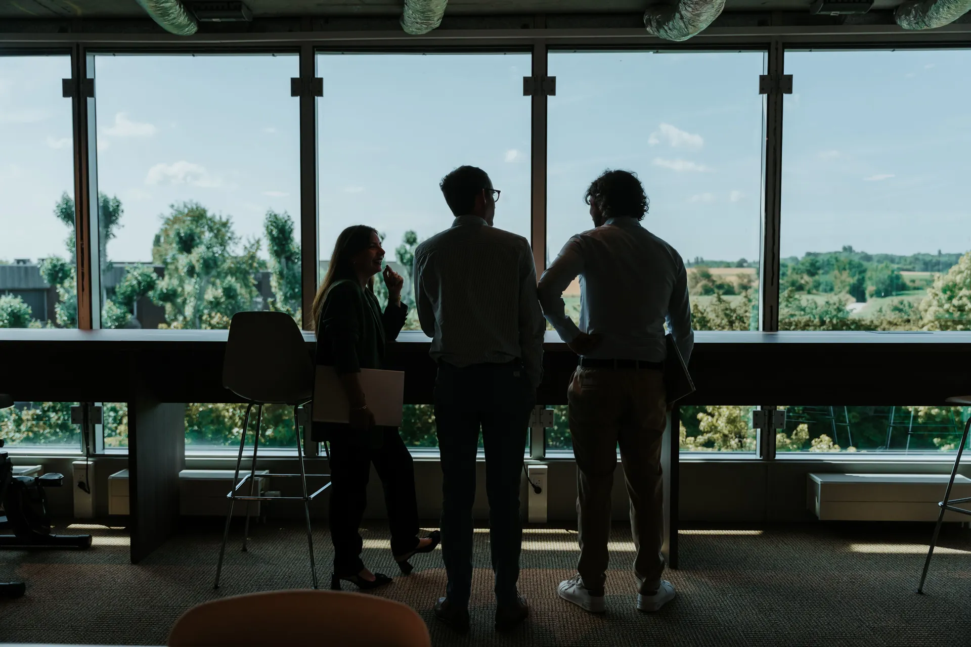 Three people stand silhouetted against large windows, discussing in an office with a scenic view of trees and a clear sky outside.