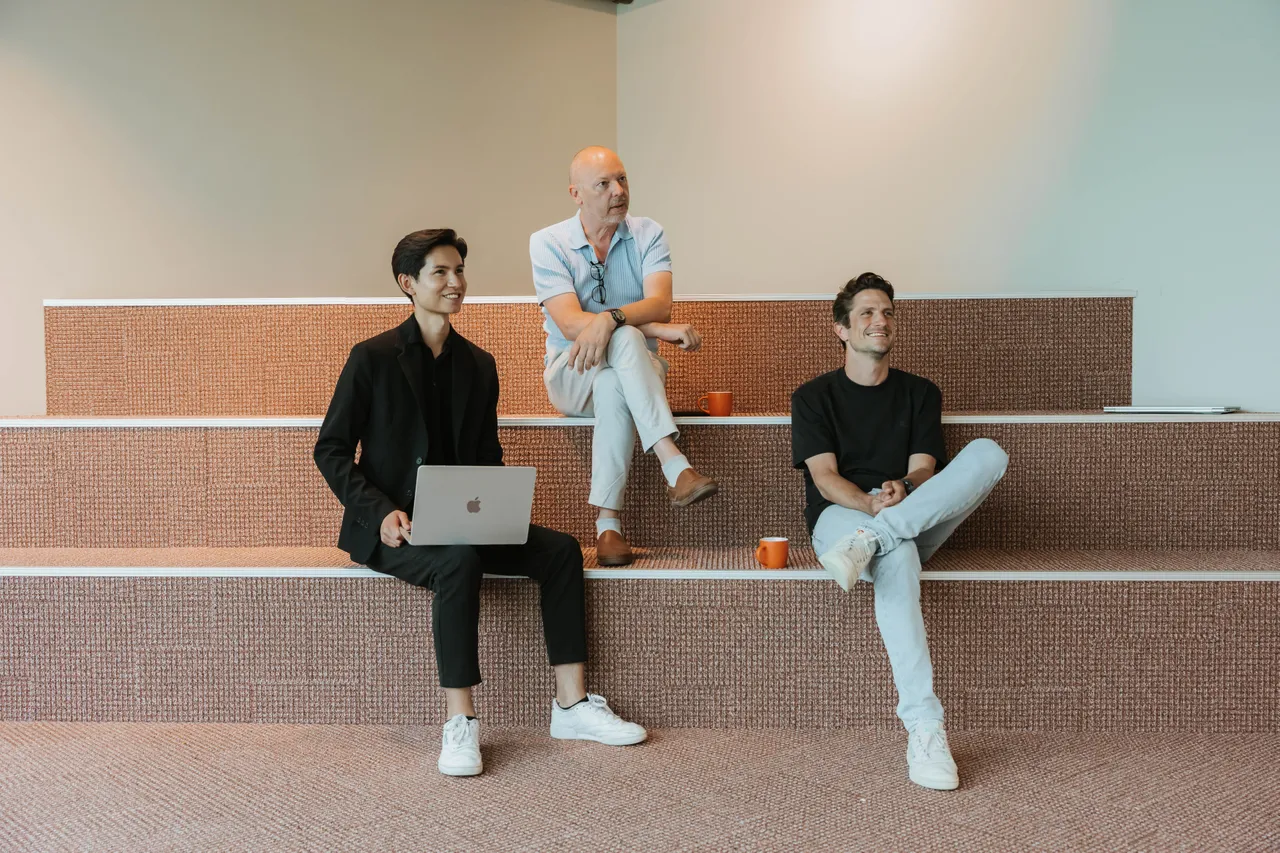 Three people sitting on tiered seating, two holding laptops, one with a coffee cup. They appear engaged and relaxed in a modern setting.
