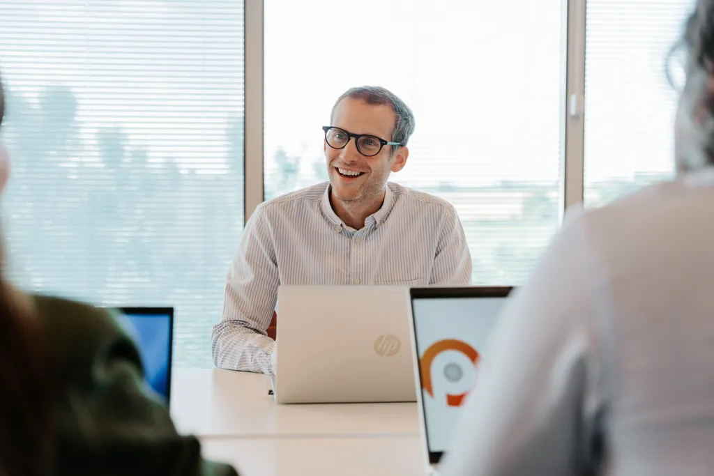 Man in glasses smiling during a meeting, sitting at a table with laptops, in a bright office with large windows.