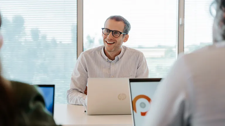 Man in glasses smiling during a meeting, sitting at a table with laptops, in a bright office with large windows.