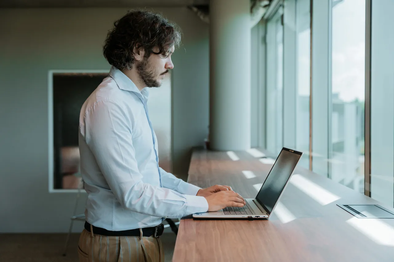 Man in a white shirt working on a laptop at a standing desk by large windows, with natural light streaming in.
