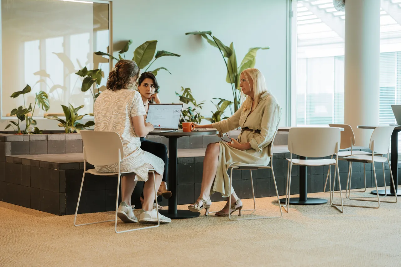 Three women sitting at a round table in a modern office space, surrounded by plants, engaged in conversation.