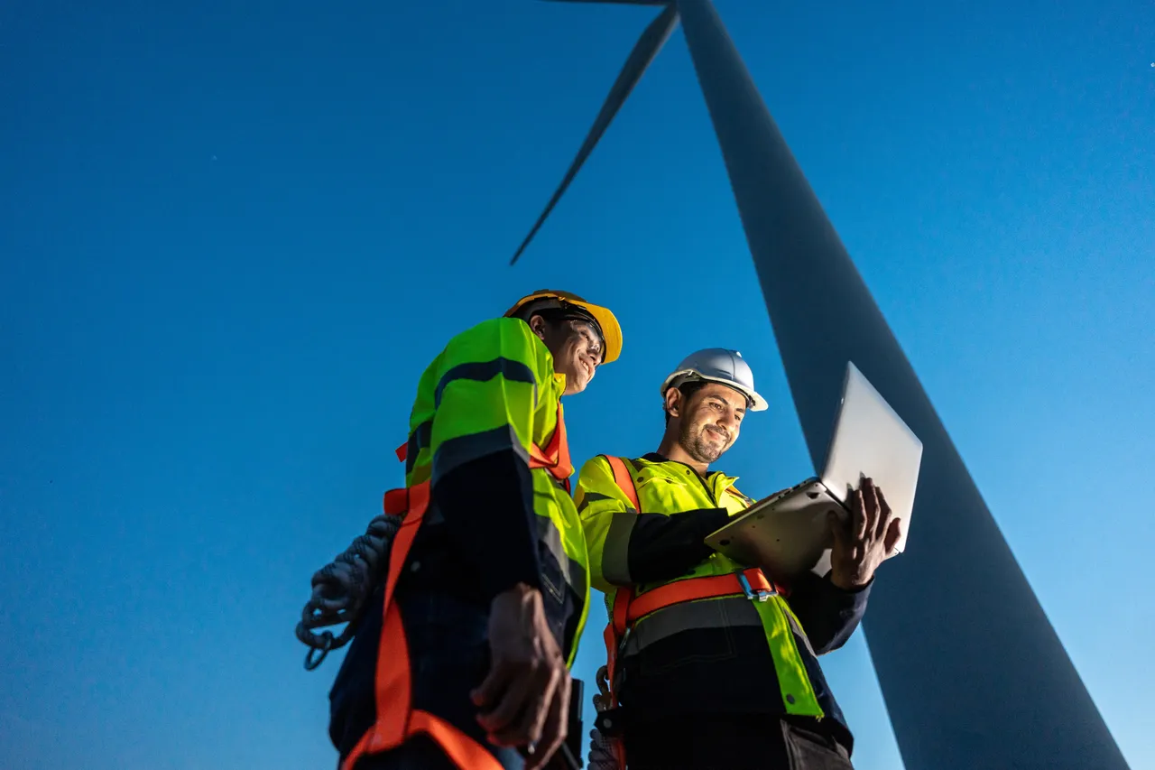 Diverse ethnicity male technicians working in the wind turbines field. Innovation engineer men use laptop computer work to develop environmental renewable clean energy outdoors at windmill power farm.