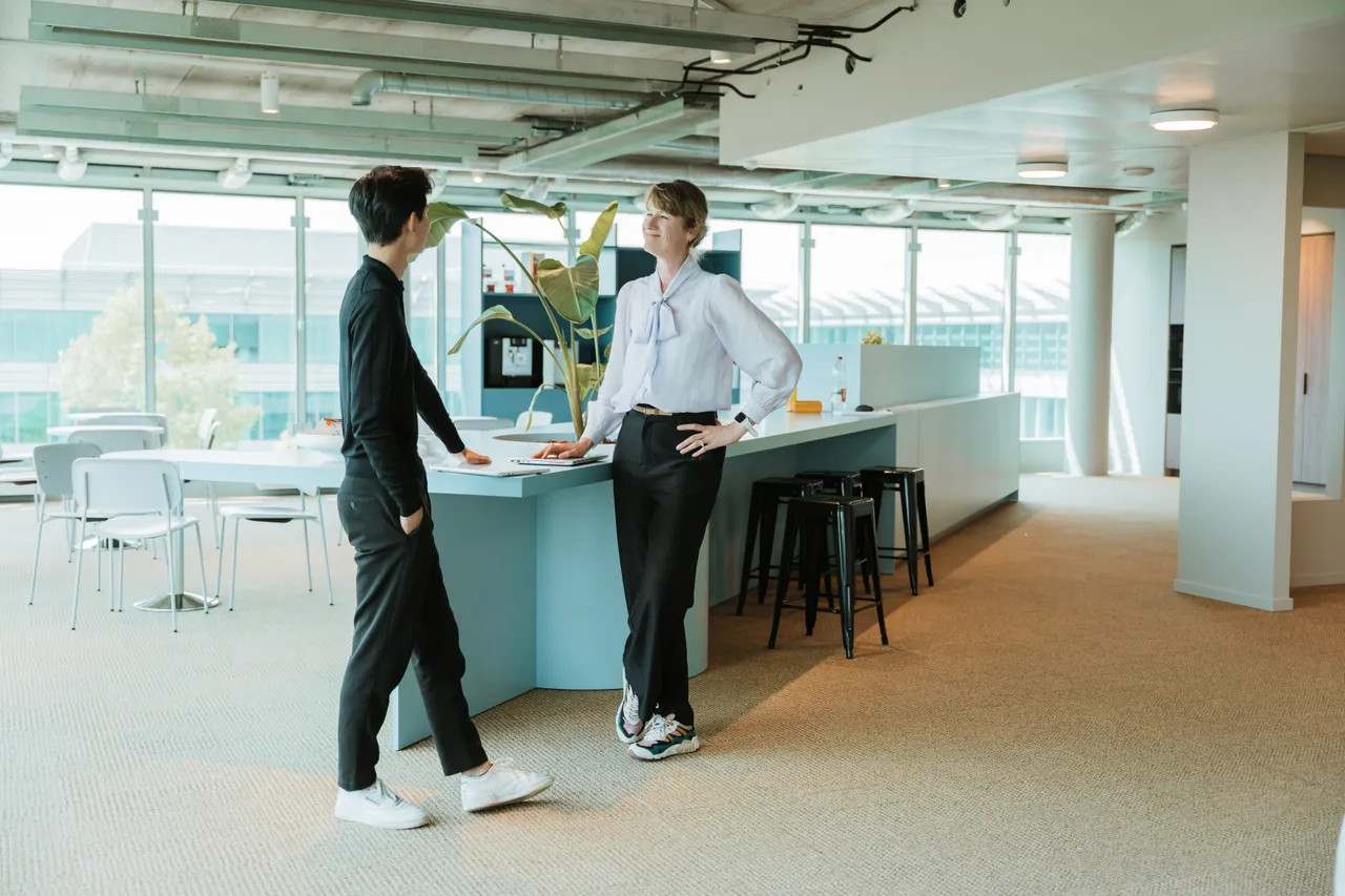 Two people conversing in a modern office space with large windows, a long table, and stools. One leans on the table, the other stands nearby.