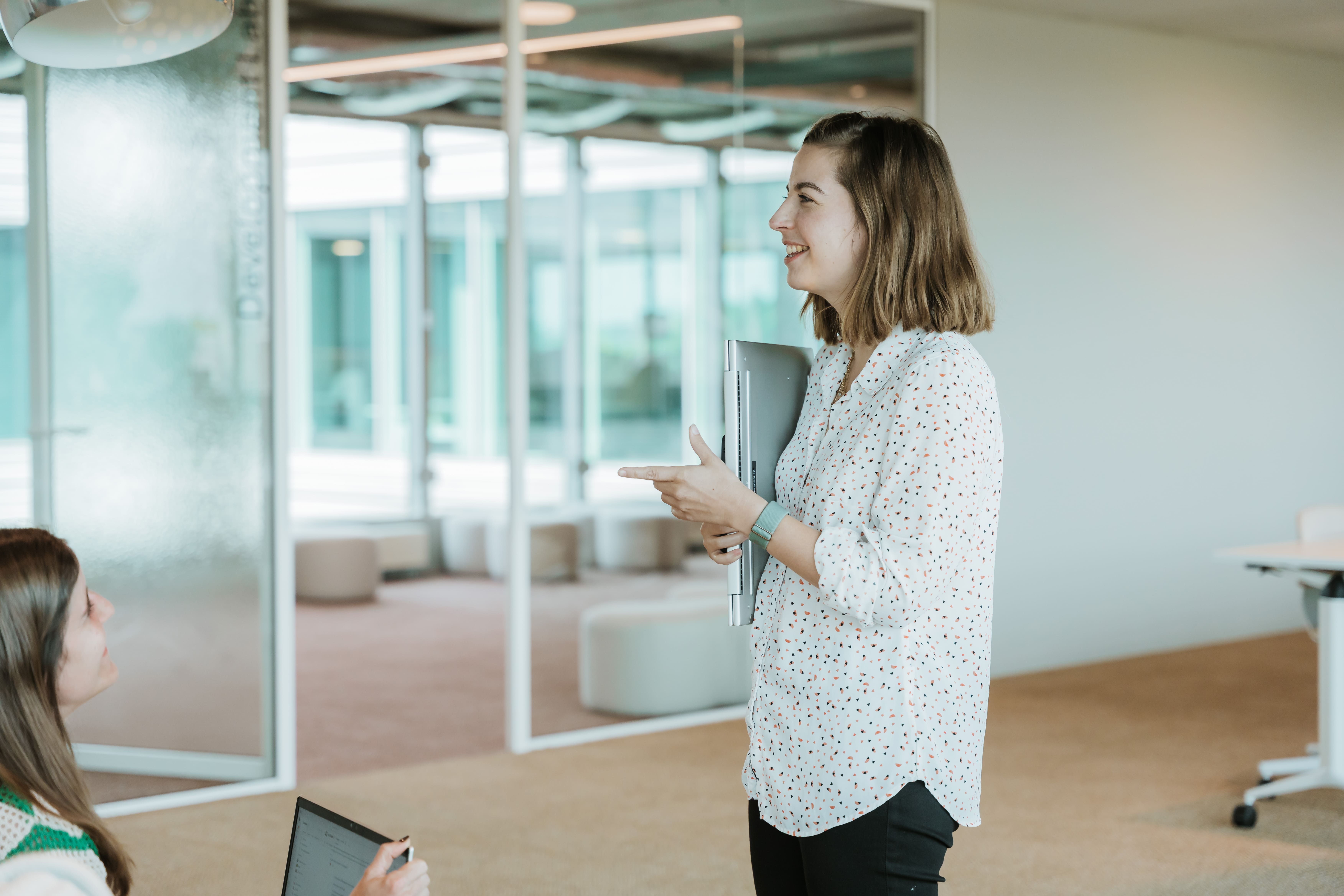 A woman in a white shirt holds a laptop and gestures while speaking to a seated colleague in a modern office space.