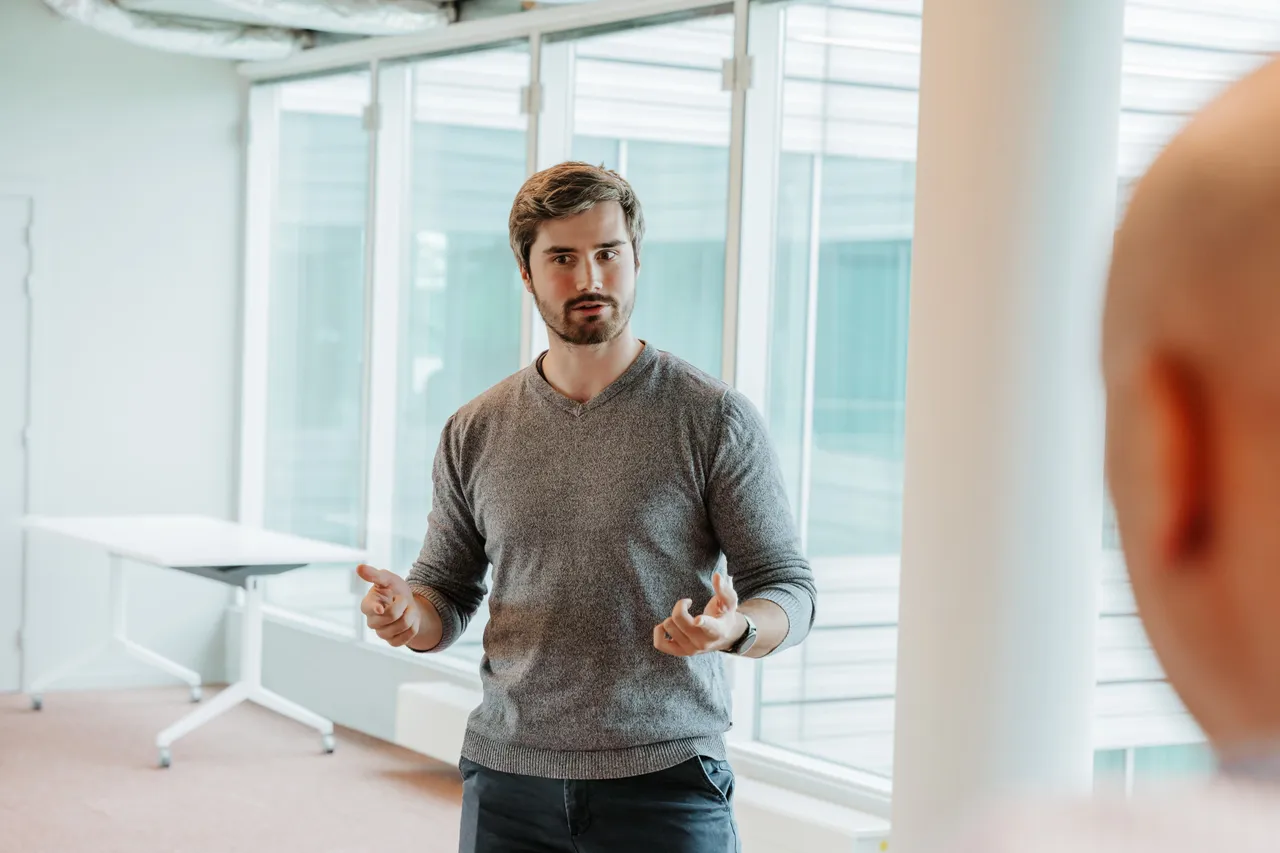 A man in a gray sweater gestures while speaking in a modern office space with large windows.