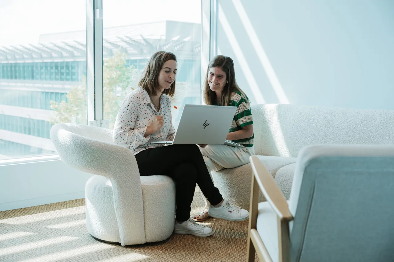 Two women sitting on a couch, looking at a laptop in a bright room with large windows and modern furniture.