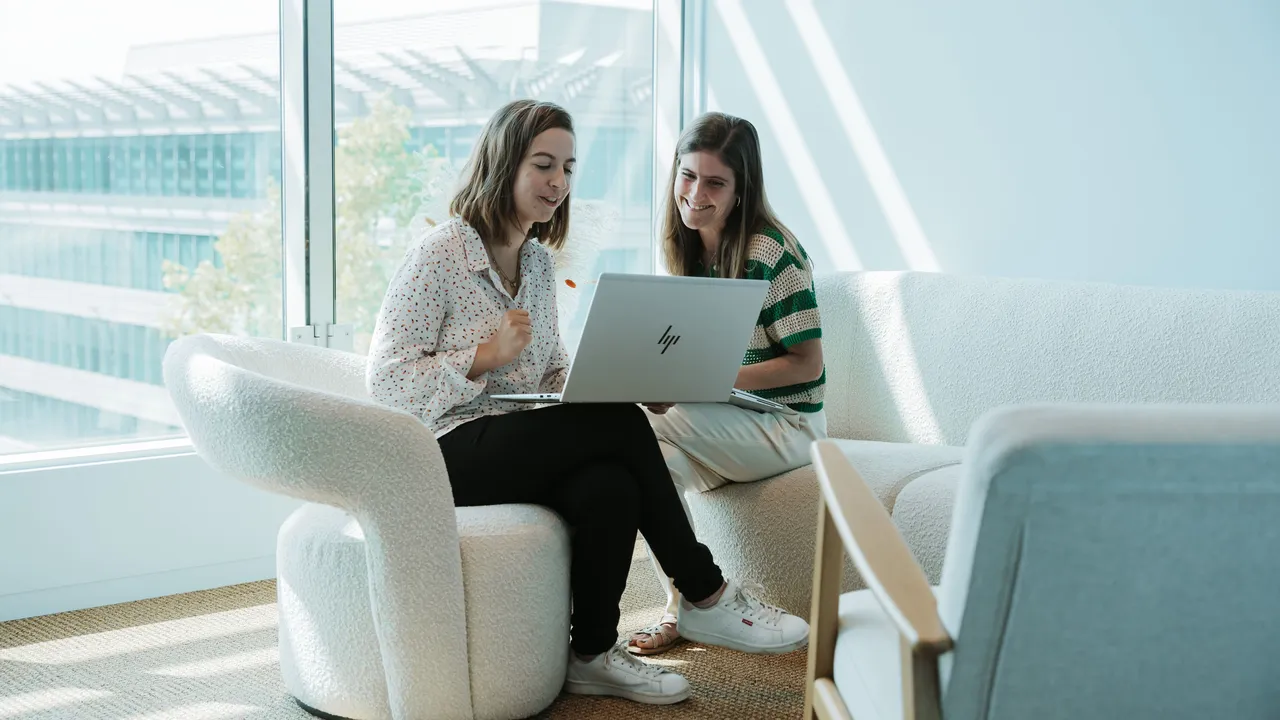 Two women sitting on a couch, looking at a laptop in a bright room with large windows and modern furniture.