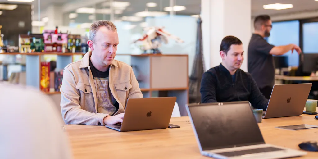 Twee mensen zitten aan een houten tafel en werken op laptops in een moderne kantooromgeving. Ze lachen en lijken in gesprek te zijn. De achtergrond toont andere werknemers, bureaus en kantoorapparatuur.