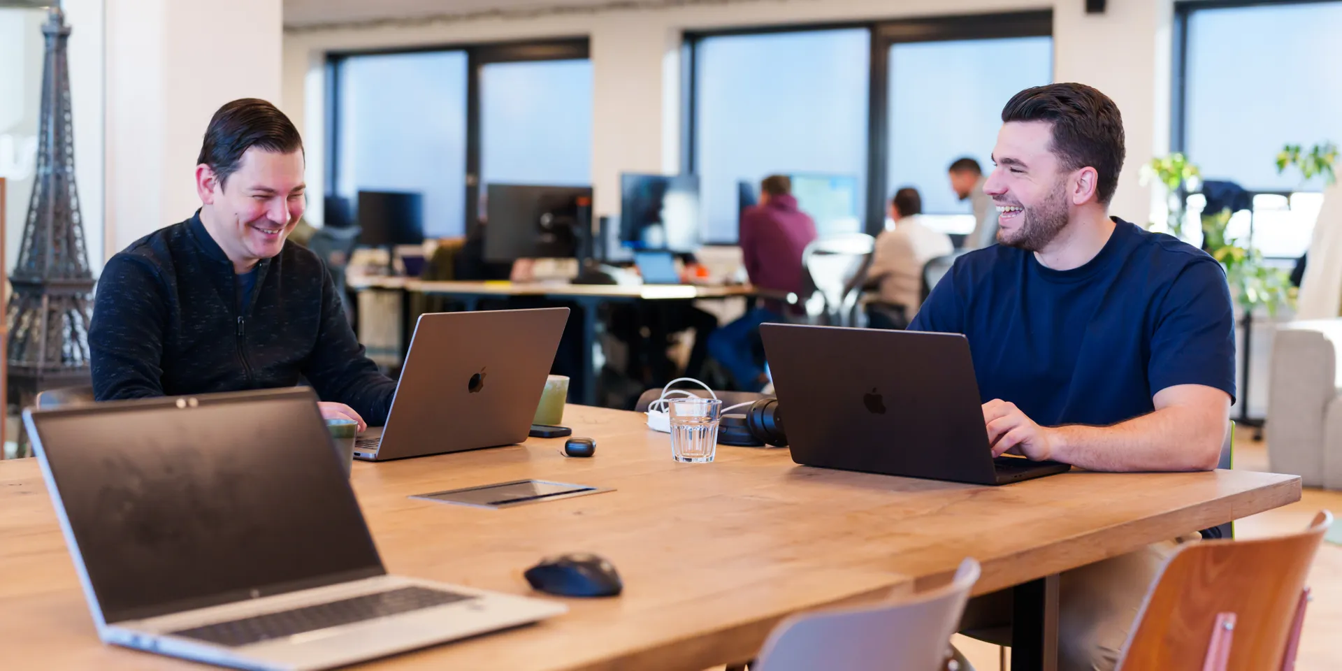 Twee mannen zitten aan een houten tafel in een kantoor, elk met een laptop. Ze lachen en zijn in gesprek. De achtergrond toont andere mensen die aan bureaus en grote ramen werken.