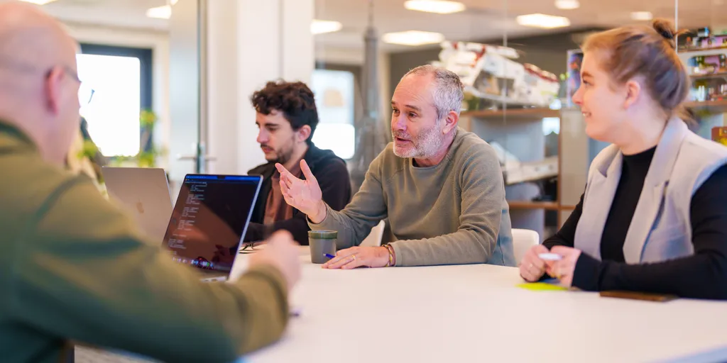 Vier mensen zitten aan een vergadertafel en zijn in gesprek. Eén persoon maakt gebaren terwijl hij spreekt en de anderen luisteren aandachtig. Laptops en notitieblokken staan op tafel. De setting is een modern kantoor met glazen wanden en planken op de achtergrond.