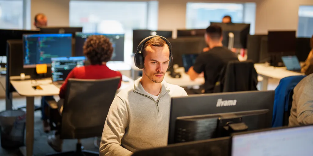 Een man met een koptelefoon is gefocust op zijn computerscherm in een moderne kantooromgeving. Achter hem werken verschillende andere mensen aan hun bureaus, met computerschermen zichtbaar. De kamer is goed verlicht met natuurlijk licht van de ramen.