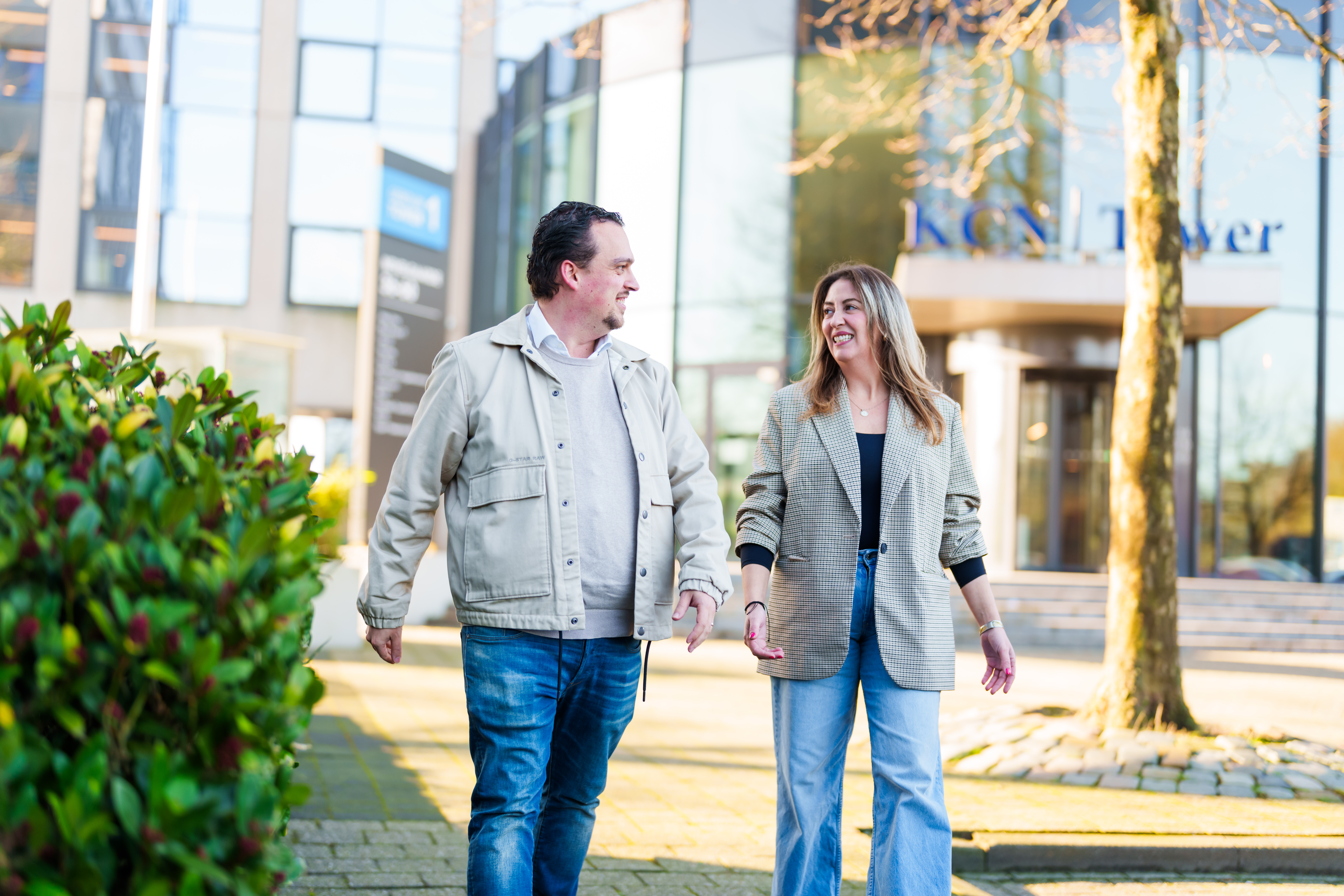 Een man en een vrouw lopen op een stoep in een stedelijk gebied. Ze lachen en lijken een gesprek te voeren. De man draagt een licht jasje en een spijkerbroek, terwijl de vrouw een geruite blazer en een spijkerbroek draagt. Op de achtergrond staat een modern gebouw.