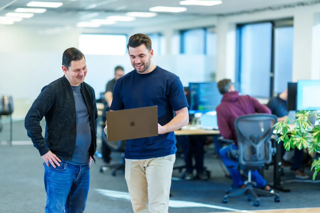 Vier mensen zitten rond een tafel in een kantooromgeving, verwikkeld in een discussie. Eén persoon gebaart terwijl hij spreekt, en twee laptops zijn zichtbaar op de tafel. Planken met items staan op de achtergrond.
