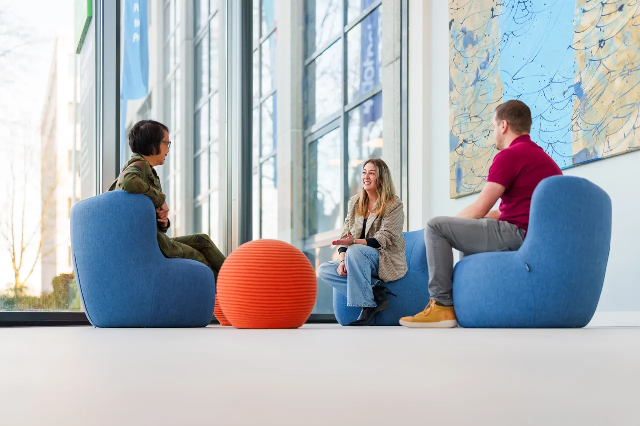Drie mensen zitten op blauwe stoelen rond een ronde oranje ottomane in een lichte, moderne kamer. Grote ramen laten natuurlijk licht binnen en een abstract schilderij siert de muur. Ze lijken in gesprek te zijn.