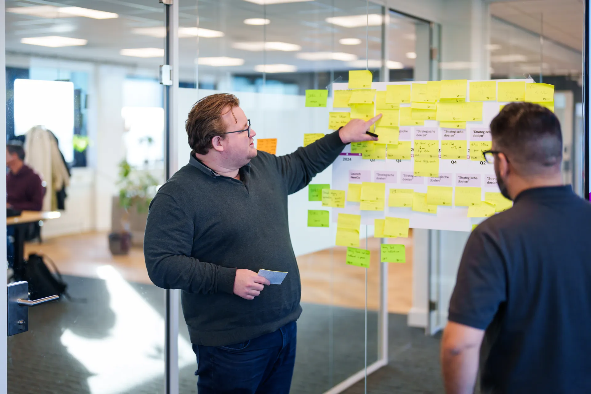 Een persoon in een trui wijst naar een bord vol met kleurrijke plakbriefjes in een modern kantoor. Een ander persoon observeert. De sfeer lijkt samenwerkend en geconcentreerd. Er zijn bureaus en mensen die op de achtergrond werken.