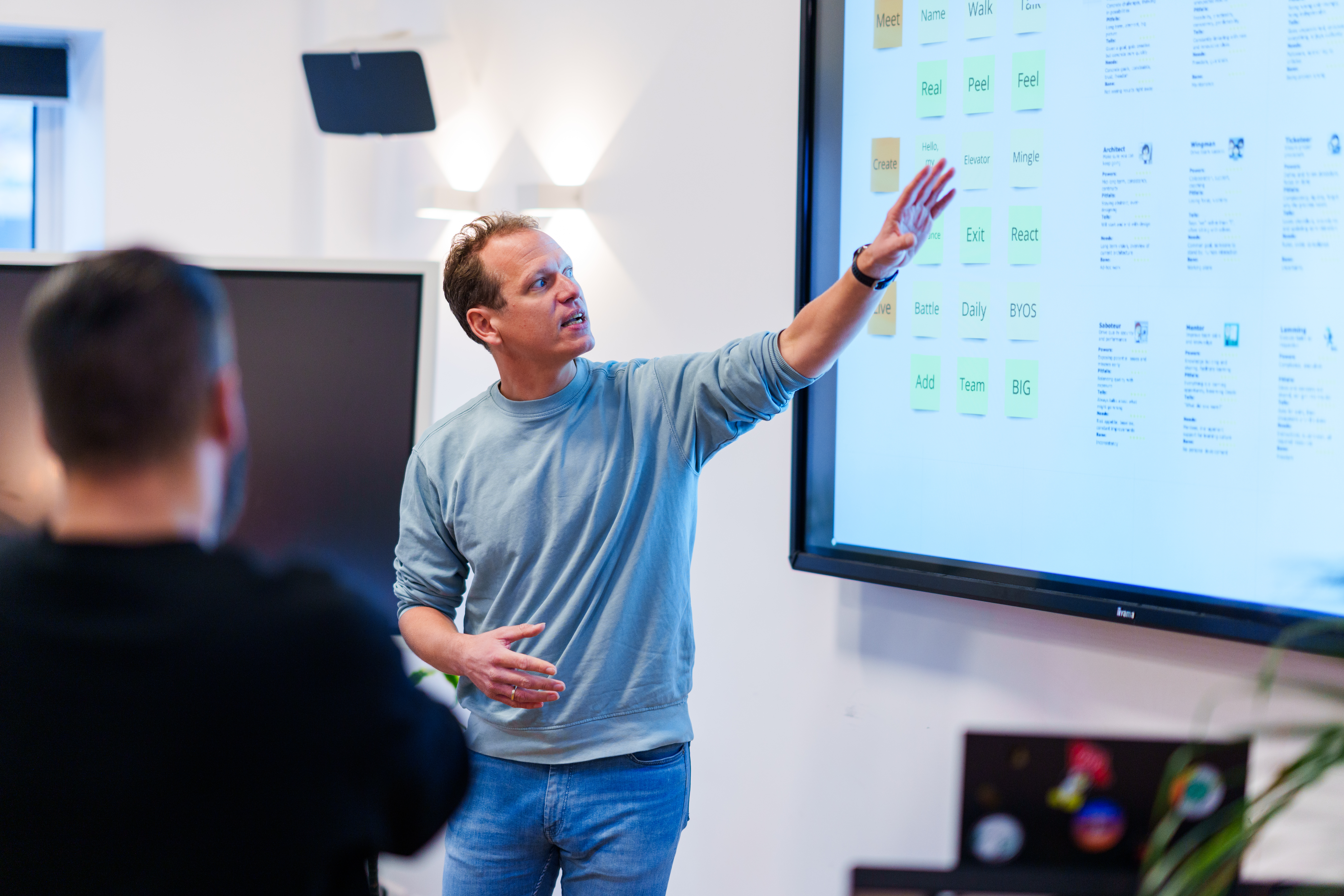 Een persoon in een blauwe trui staat en wijst naar een groot scherm aan de muur met kleurrijke plaknotities. Een andere persoon, wazig op de voorgrond, kijkt naar het scherm. De setting lijkt een kantoor of vergaderruimte te zijn.