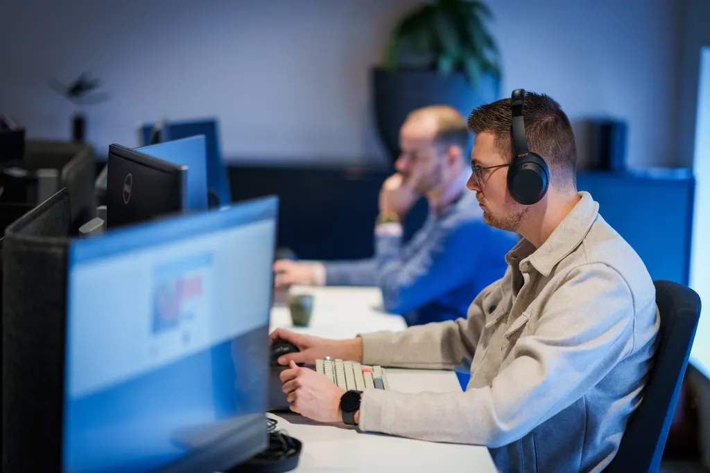 Een persoon met een koptelefoon is gefocust op het werken op een computer op kantoor. Een andere persoon op de achtergrond werkt ook op een computer, gedeeltelijk wazig. Een plant is zichtbaar op een plank op de achtergrond.