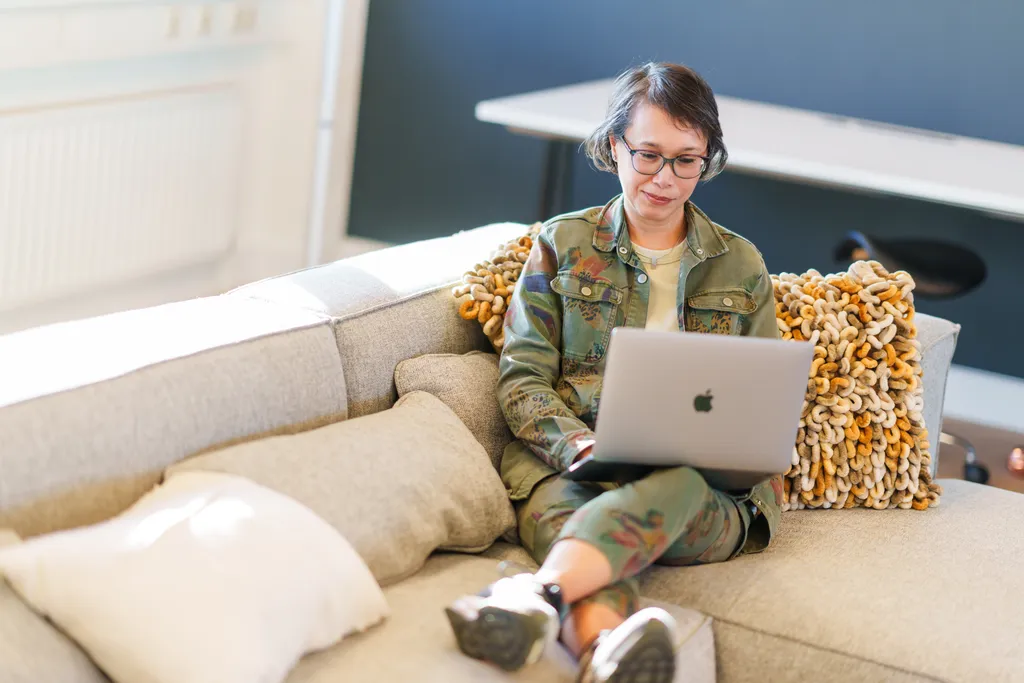 Een persoon met kort haar en een bril zit op een grijze bank en gebruikt een zilveren laptop. Ze dragen een groene camouflage-outfit. Een getextureerde mosterdgele deken is over de bank gedrapeerd. Op de achtergrond is een bureau zichtbaar.