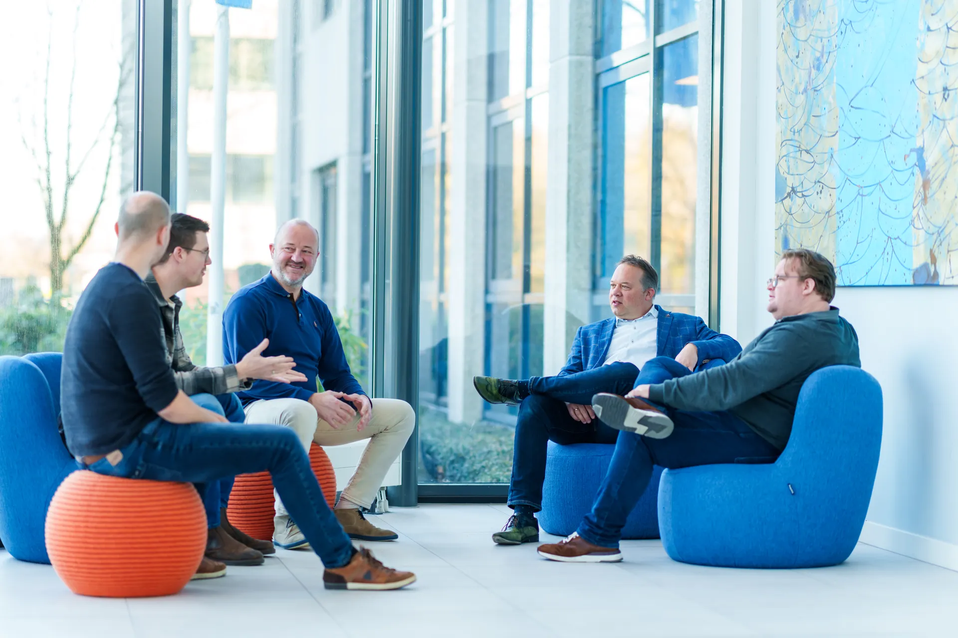 Vijf mannen zitten in een moderne kantoorlounge. Twee zitten op oranje, ronde krukken en drie op gebogen blauwe stoelen. Ze zijn in gesprek, met een groot raam en een decoratief muurstuk op de achtergrond.