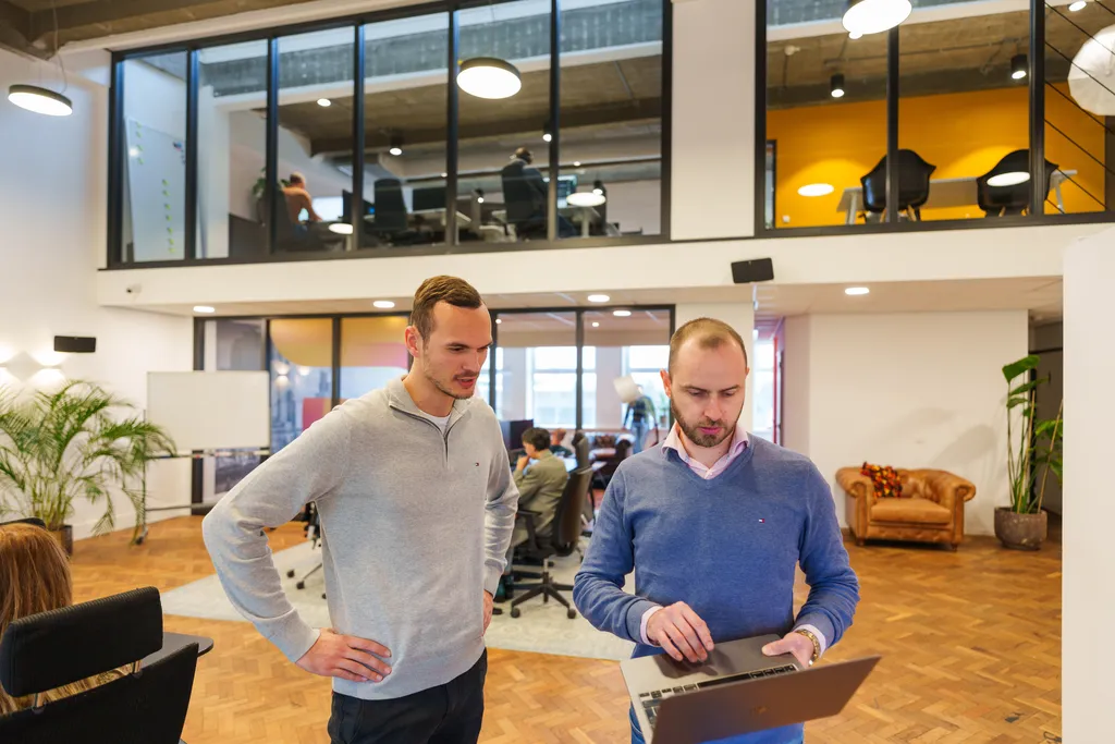 Twee mannen kijken naar een laptop in een moderne kantoorruimte met een houten vloer. De een draagt een lichtgrijze trui, de ander een blauwe. Op de achtergrond werken mensen aan bureaus en is een open mezzanine zichtbaar.