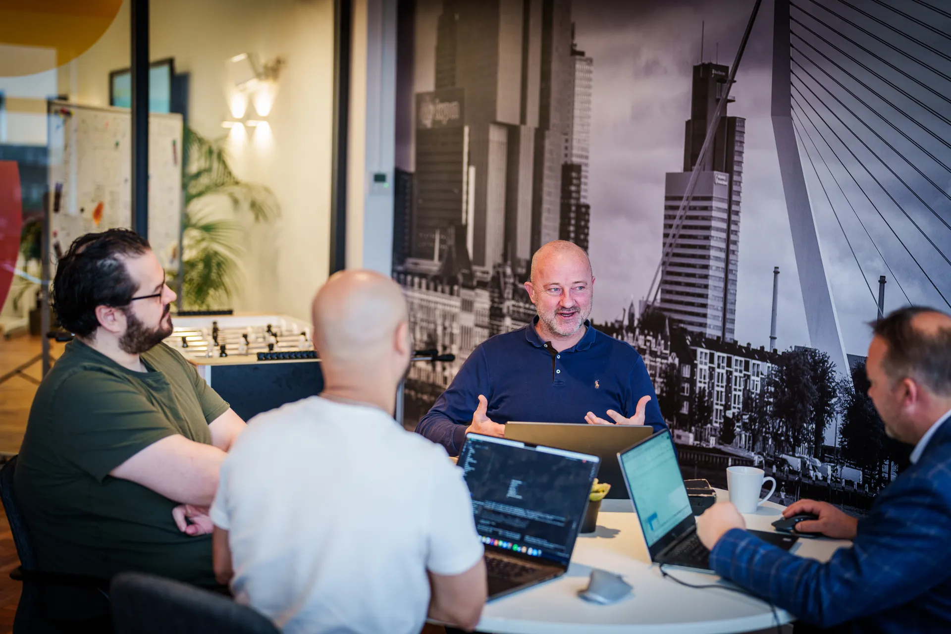 Vier mensen discussiëren rond een tafel met open laptops. Eén persoon maakt gebaren terwijl hij spreekt. Op de achtergrond staat een grote zwart-witte muurschildering van een stadsgezicht.