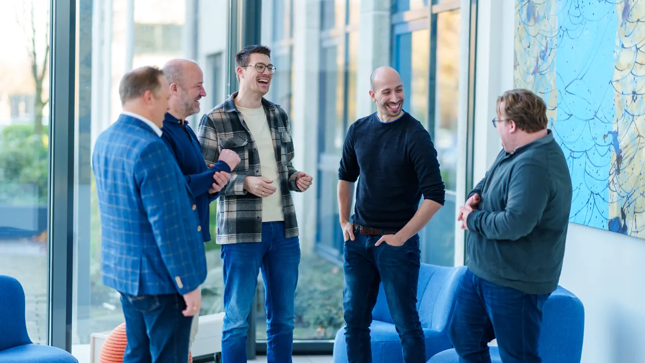 Vijf mannen in casual kleding staan in een lichte, moderne kamer met grote ramen en blauwe stoelen. Ze zijn in een vriendelijk gesprek verwikkeld, lachend en gebaren makend.