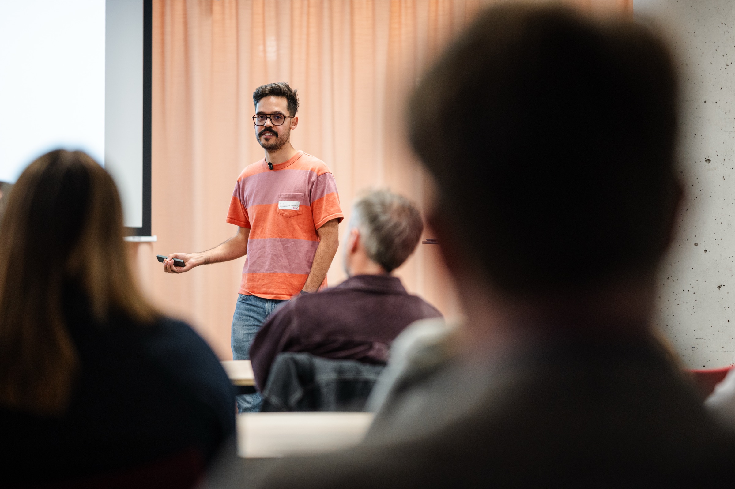 A speaker in a striped T-shirt addresses the room during the first session, seen past the heads of attendees.